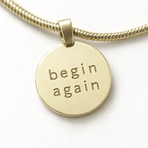Close-up of the Origin Disc pendant showing begin again engraved into the gold, with its tube bail and snake chain