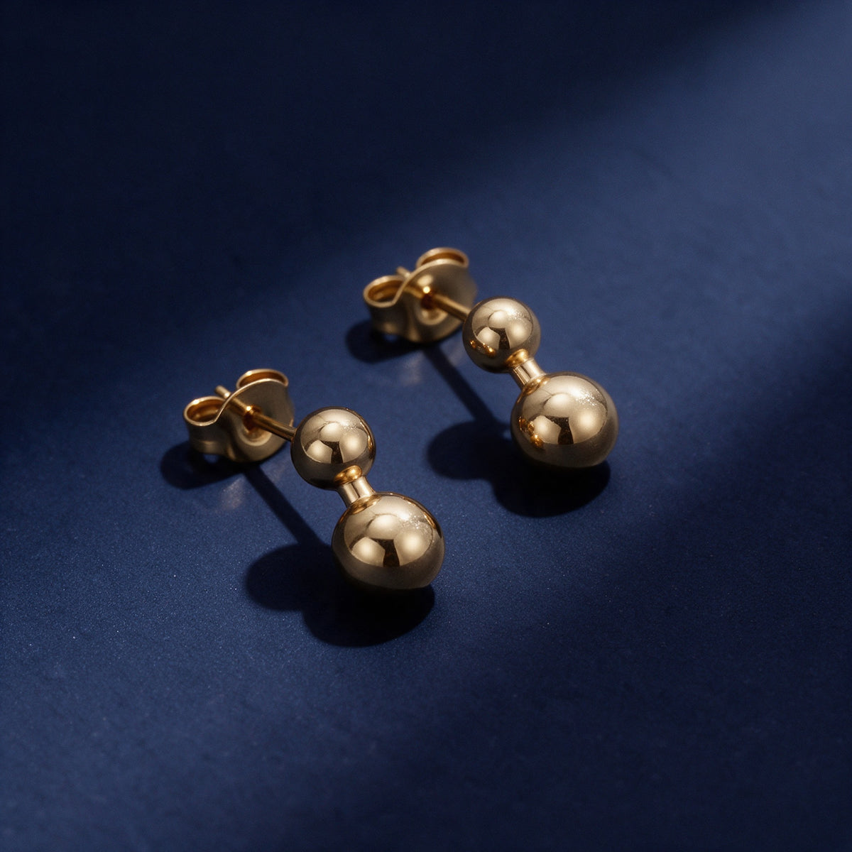 Echo Studs, a pair of gold barbell stud earrings on deep navy