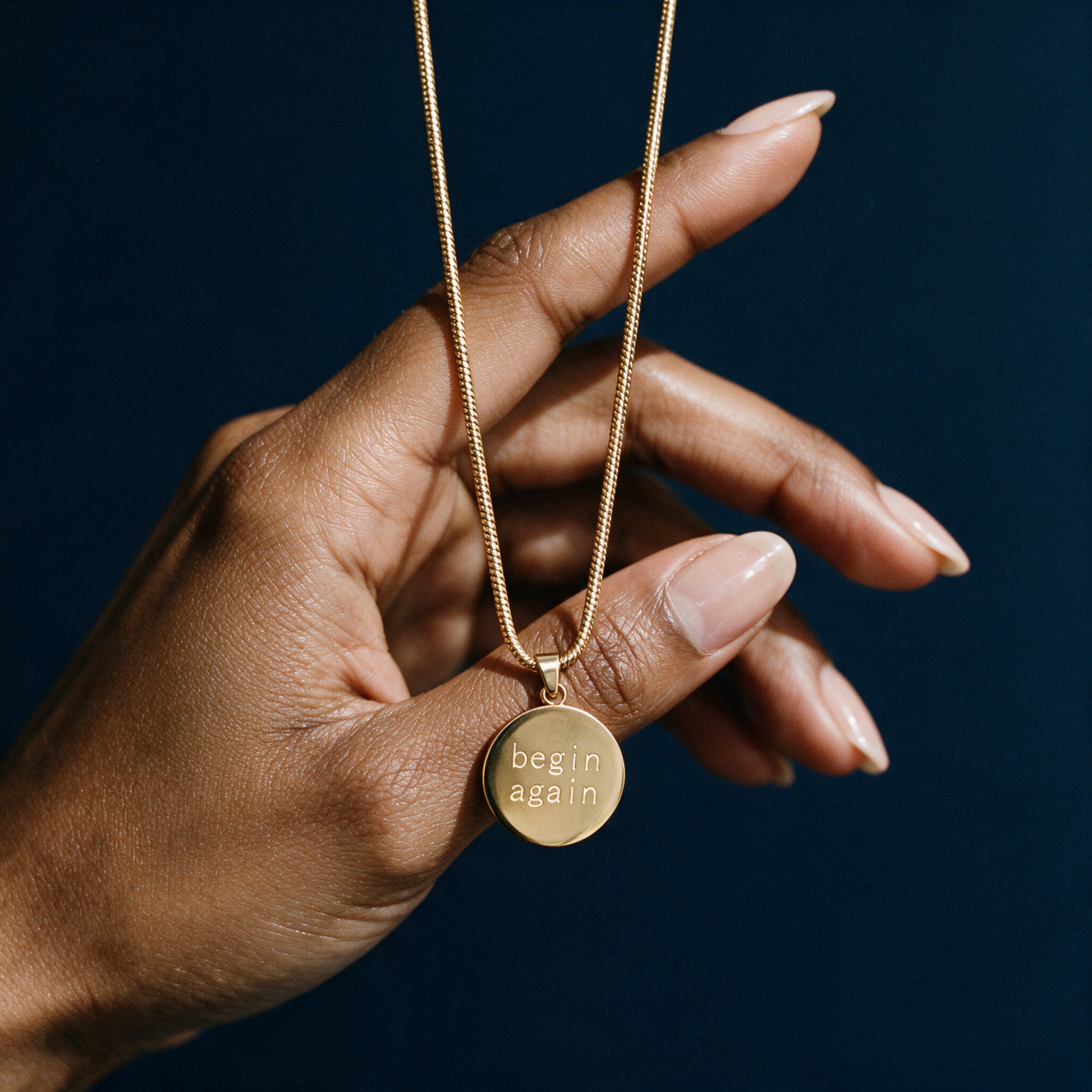 Origin Disc necklace engraved begin again on a fine gold chain, held in a hand against a deep navy background
