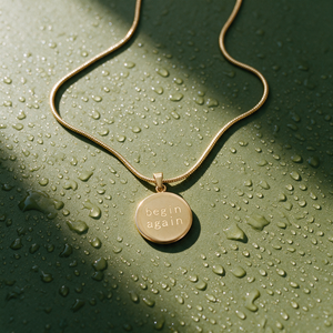 Origin Disc necklace engraved begin again on a fine gold chain, resting on an olive surface scattered with water droplets