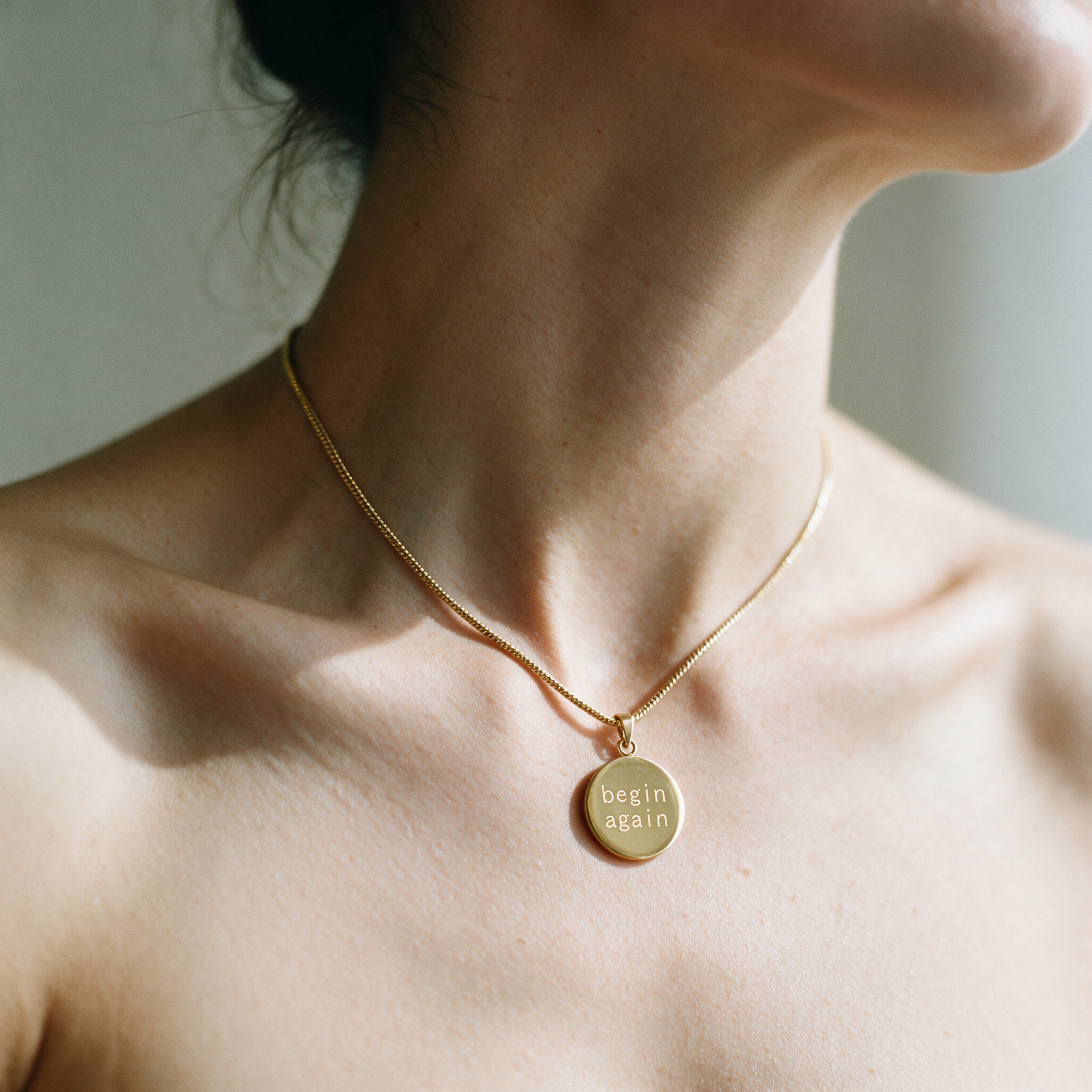 Origin Disc necklace worn on the neck, a fine gold chain with a round pendant engraved begin again